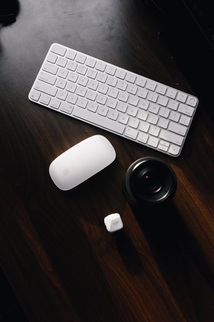 Flat lay of a wireless keyboard, mouse, camera lens, and black table creating a stylish workspace.